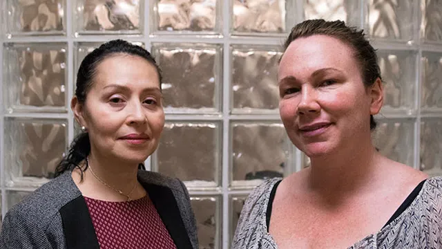 Two women standing indoors in front of glass block wall, both looking at the camera with neutral expressions.