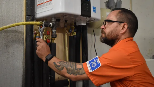 A technician adjusting plumbing connections on a water heater, wearing an orange uniform and safety glasses.