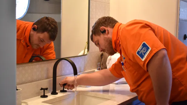 A technician in an orange shirt installs a modern sink faucet in a stylish bathroom with a mirror.