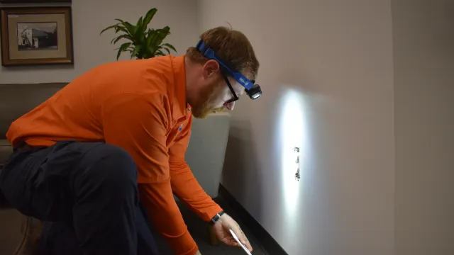 Technician in orange shirt wearing headlamp fixes electrical outlet on wall in indoor room with carpet.