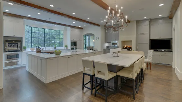 Spacious modern kitchen with large marble island, wood beams, chandelier, and built-in appliances against white cabinetry.