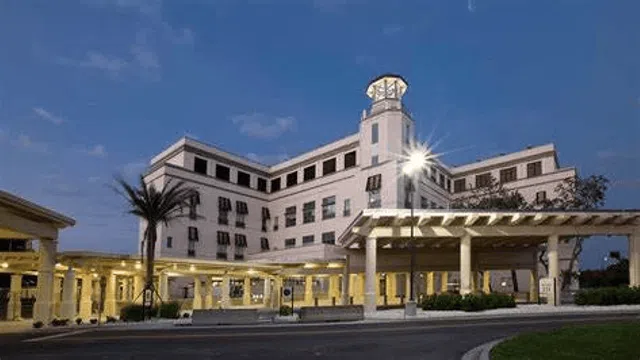 Exterior of a large, white hospital building with palm trees and covered entrance at dusk with lights on
