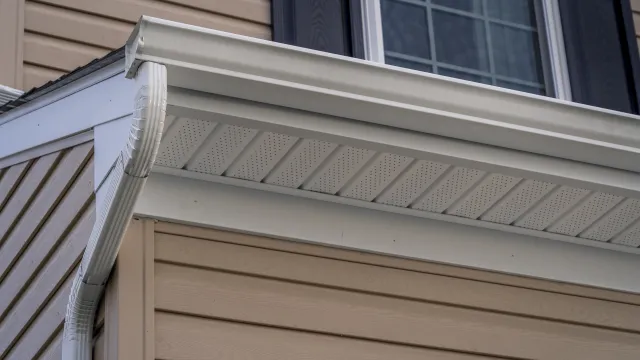 White vinyl rain gutter and downspout installed on beige house exterior with window and black shutters.