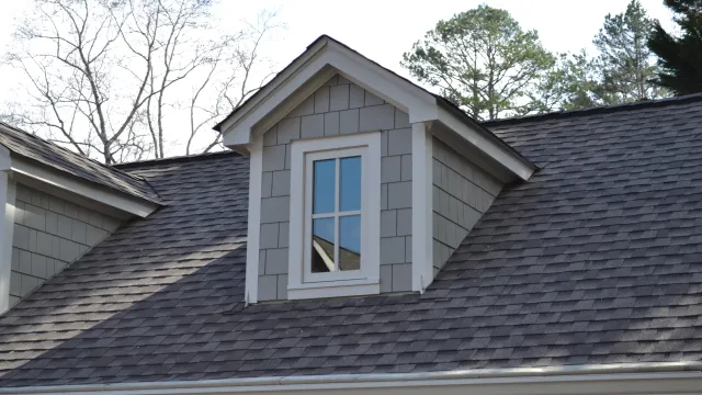 Gray shingled dormer window on a dark gray asphalt roof with trees in the background