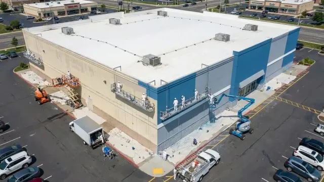 Workers repainting the exterior of a large commercial building with blue and white colors using lifts and scaffolding.