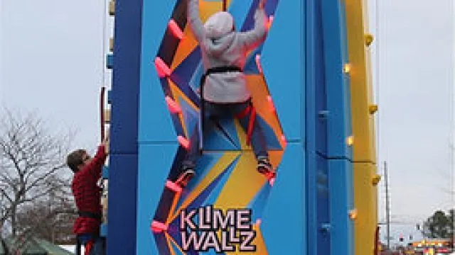 Child climbing an outdoor colorful climbing wall named Klime Wallz with people watching nearby at an event.