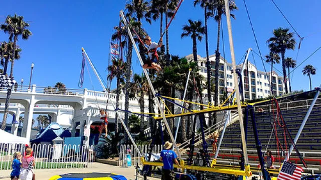Children bouncing on trampolines and using bungee cords in a sunny park with palm trees and a white arch bridge.