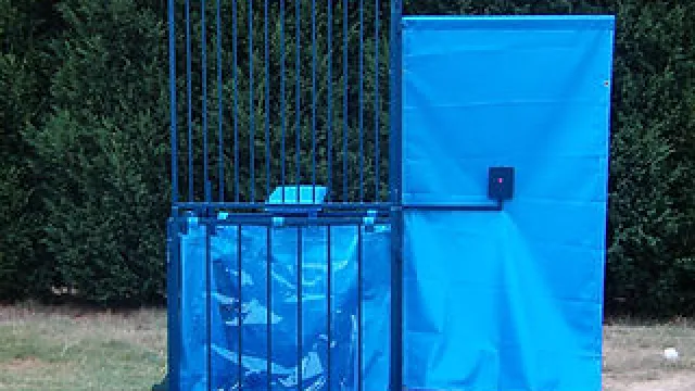 Blue dunk tank with metal cage and splash panel set outdoors on grass near green hedge.