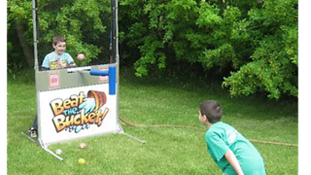 Two boys play the Beat the Bucket ball tossing game outdoors on green grass near trees.