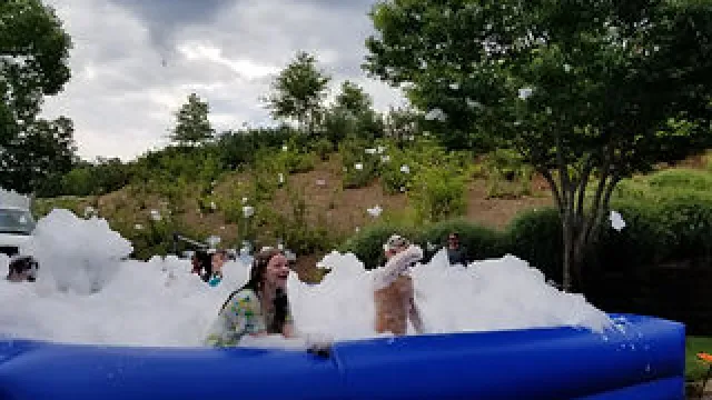 People enjoying a foam party in a blue inflatable pool outdoors surrounded by trees and plants.