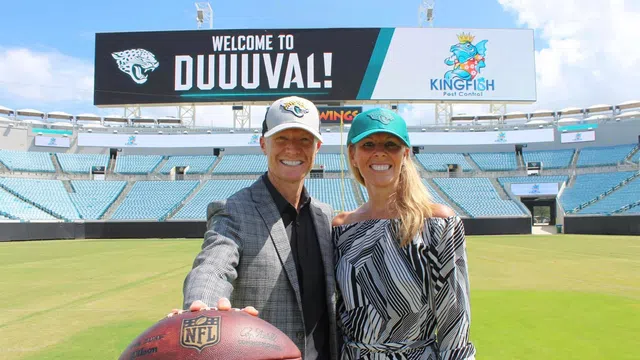Smiling man and woman in Jacksonville Jaguars caps pose on football field holding NFL ball under 'Welcome to DUUUVAL' sign.