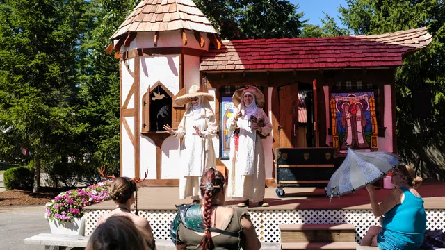 Performers in white robes acting on stage in front of a medieval-themed set with audience watching outdoors.