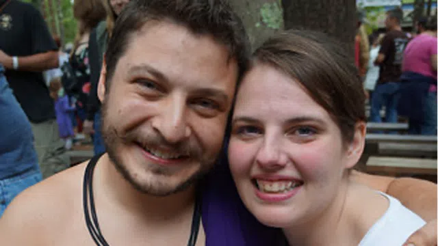 Couple smiling happily, man holding open ring box, woman holding red rose, outdoor gathering in the background
