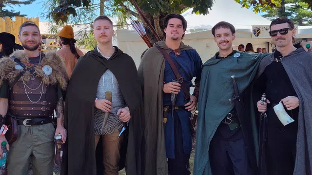 Five men dressed in medieval costumes with cloaks and weapons posing outdoors in daylight under trees.