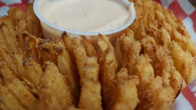 Crispy fried blooming onion served with a creamy dipping sauce on red and white checkered paper.