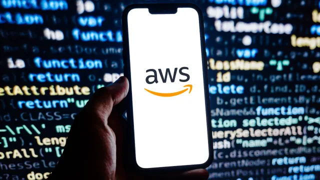 Hand holding phone displaying AWS logo with blurred programming code in background representing cloud computing.
