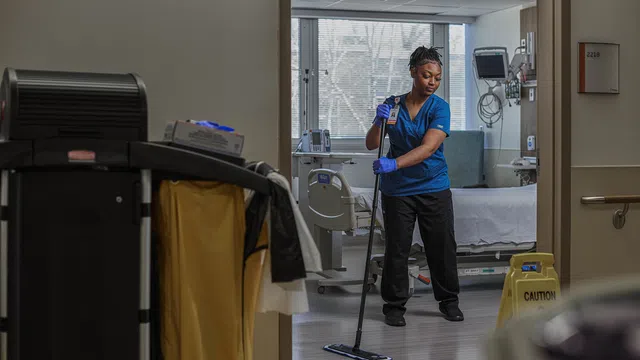 Healthcare worker cleaning hospital room floor wearing gloves and uniform with caution sign nearby