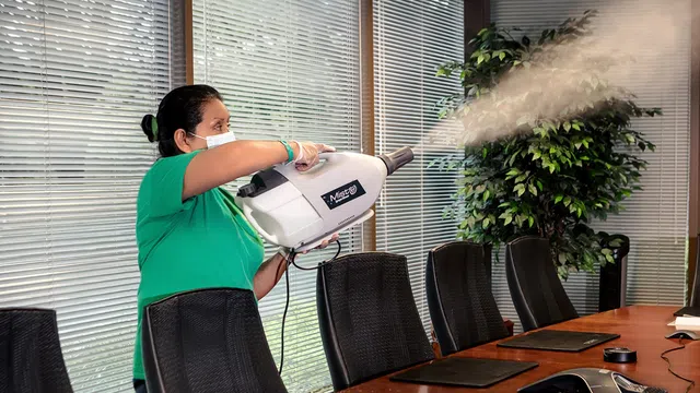 Person wearing mask spraying disinfectant fog in a modern office conference room with glass windows and chairs.