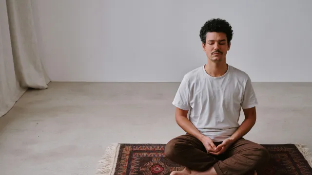 Man meditating cross-legged on patterned rug in a minimalist room with neutral tones and natural light.