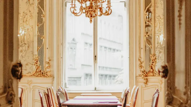 Elegant dining room with ornate chandelier, large window, and classic wooden table and chairs in golden decor.