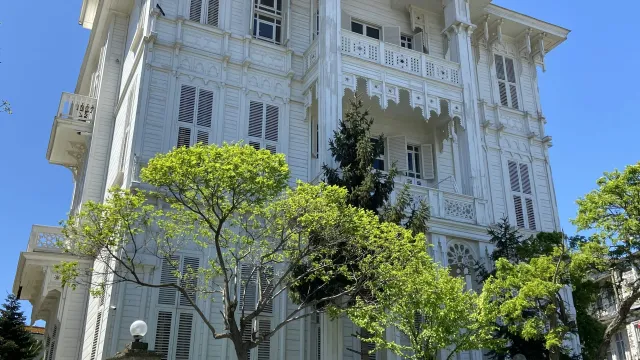 Historic white wooden mansion with ornate architectural details surrounded by green trees and clear blue sky.