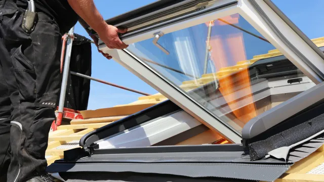 A worker installing a skylight on a rooftop under a clear blue sky.