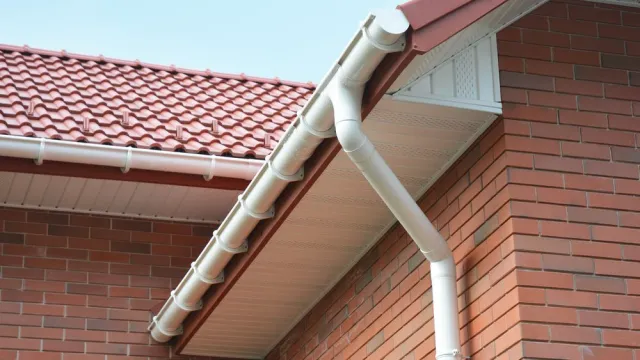 Close-up of a white gutter system installed on a red-tiled roof of a brick house.