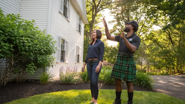 Home inspector in kilt explaining features of a house to a female client outdoors in a sunny garden.
