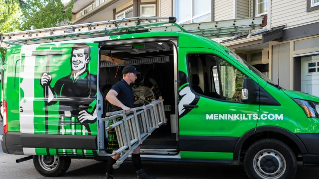 Worker carrying ladder from green Men In Kilts service van parked in front of residential building.