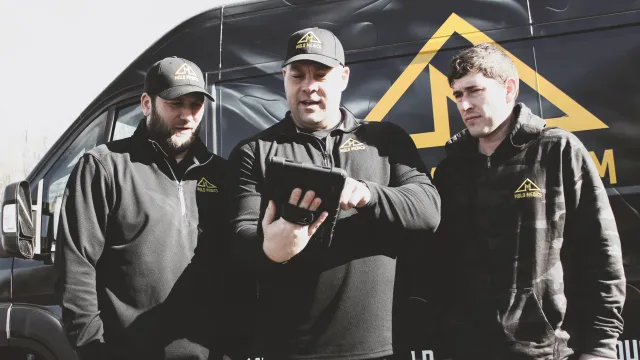 Three workers in black Milo Metrics uniforms checking a tablet beside a company van with a yellow logo.