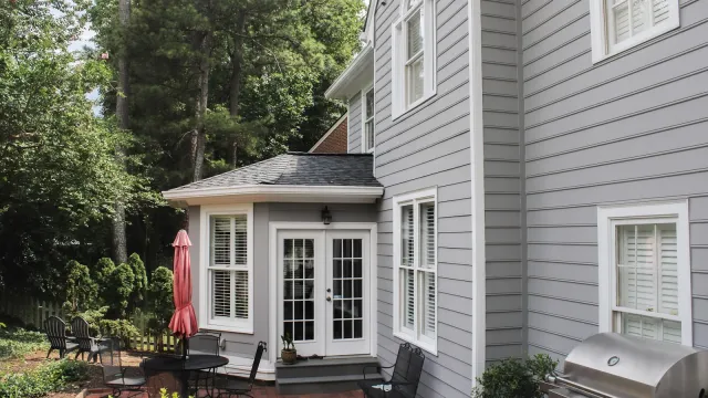 Modern two-story house with grey siding, multiple windows, wooden deck, and outdoor grill area on a sunny day.