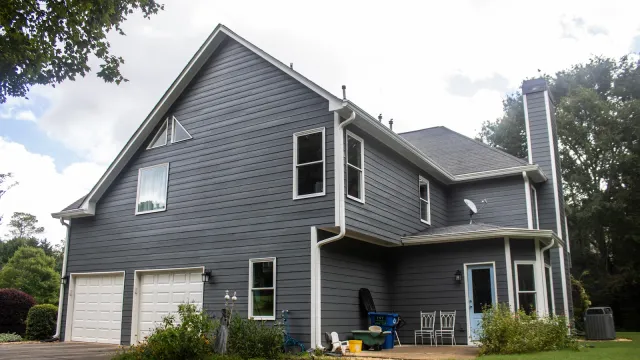 Two-story gray house with white trim, double garage, patio furniture, and surrounding greenery under a cloudy sky