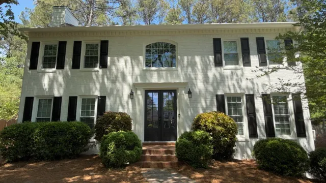 White two-story house with black shutters, glass front door, surrounded by green bushes and tall pine trees.