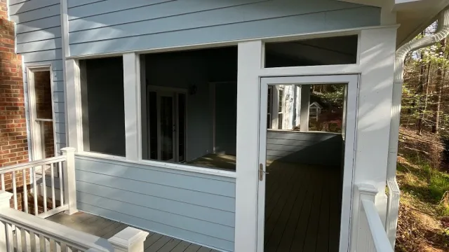 Gray screened porch with white door and railings attached to a house under clear blue sky.