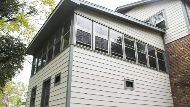 White two-story house with a glassed-in sunroom and gray trim surrounded by green trees.
