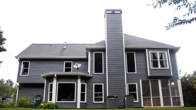 Back view of a modern two-story gray house with multiple windows, chimney, and green lawn under overcast sky
