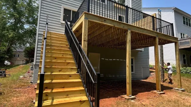 New wooden deck with stairs and black metal railing attached to a two-story gray house on a sunny day.