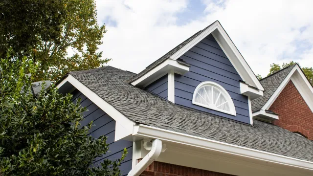 Close-up of a house roof with gray shingles, blue siding, white trim, and an arched window under a cloudy sky.