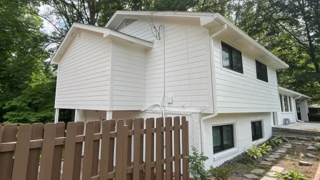 Two-story white house with black windows, surrounded by trees and a brown wooden fence in the front yard.