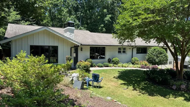 Bright ranch-style house with white brick exterior, large windows, lush green lawn, and landscaped garden under blue sky.