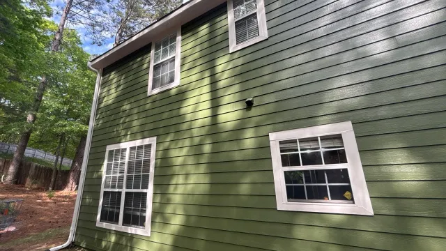 White two-story house with black shutters, glass front door, surrounded by green bushes and tall pine trees.