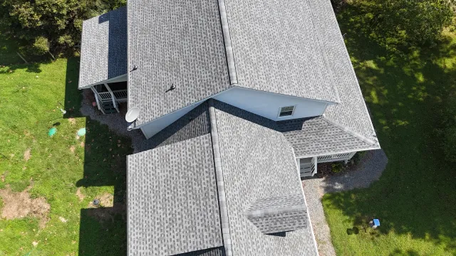 Aerial view of a grey shingled roof on a white house surrounded by green grass and trees on a sunny day.