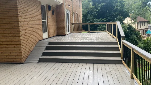 Spacious gray wooden deck with stairs and railing attached to a brick two-story house surrounded by trees.