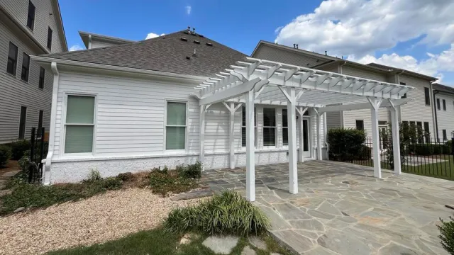 White pergola on stone patio attached to a modern white house with blue sky and scattered clouds.