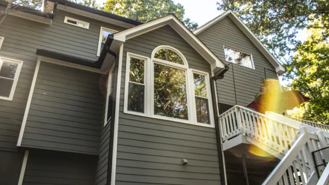 Modern two-story house with grey siding, multiple windows, wooden deck, and outdoor grill area on a sunny day.