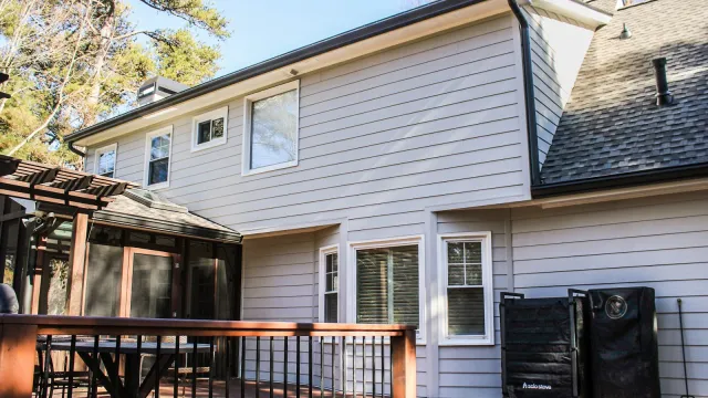 Modern two-story house with grey siding, multiple windows, wooden deck, and outdoor grill area on a sunny day.