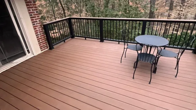 Spacious wooden deck with black metal railing and a round metal table with three chairs overlooking a wooded area.