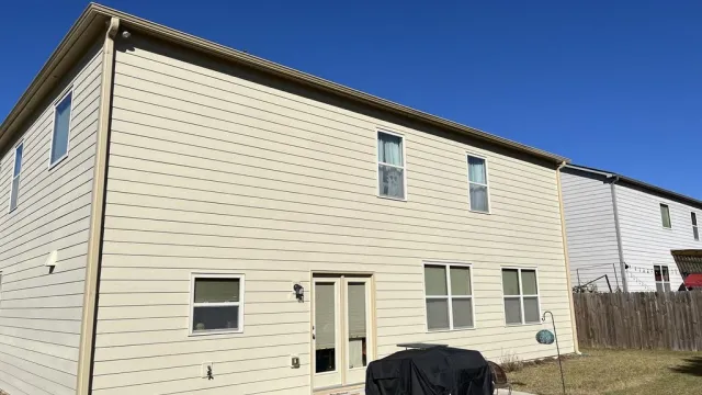 Two-story beige house with multiple windows and a backyard patio with a covered grill under clear blue sky.