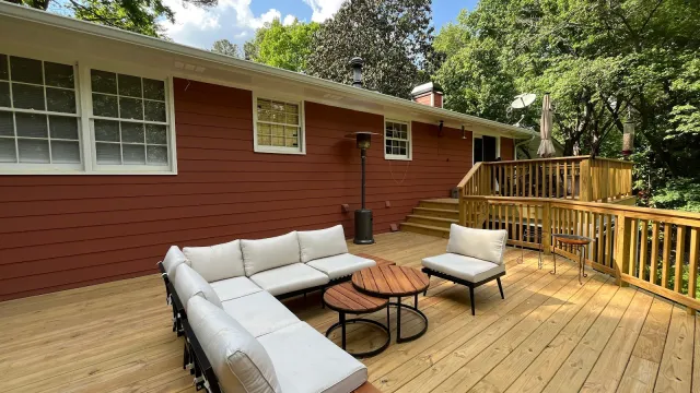 Spacious wooden deck with white cushioned sectional sofa, coffee tables, and surrounding greenery under blue sky.