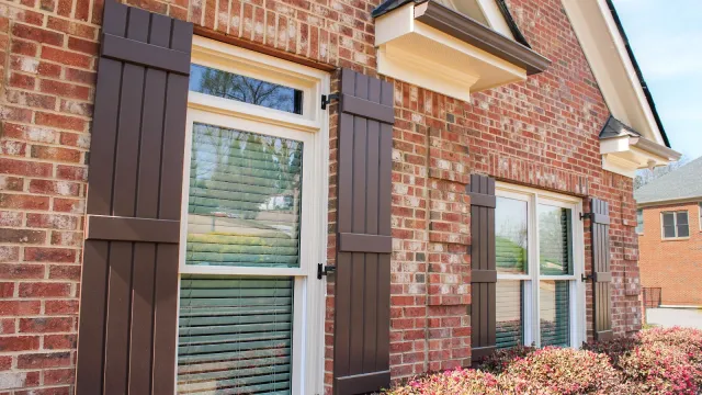 Red brick house exterior with white framed windows, dark brown shutters, and flowering bushes under bright daylight.